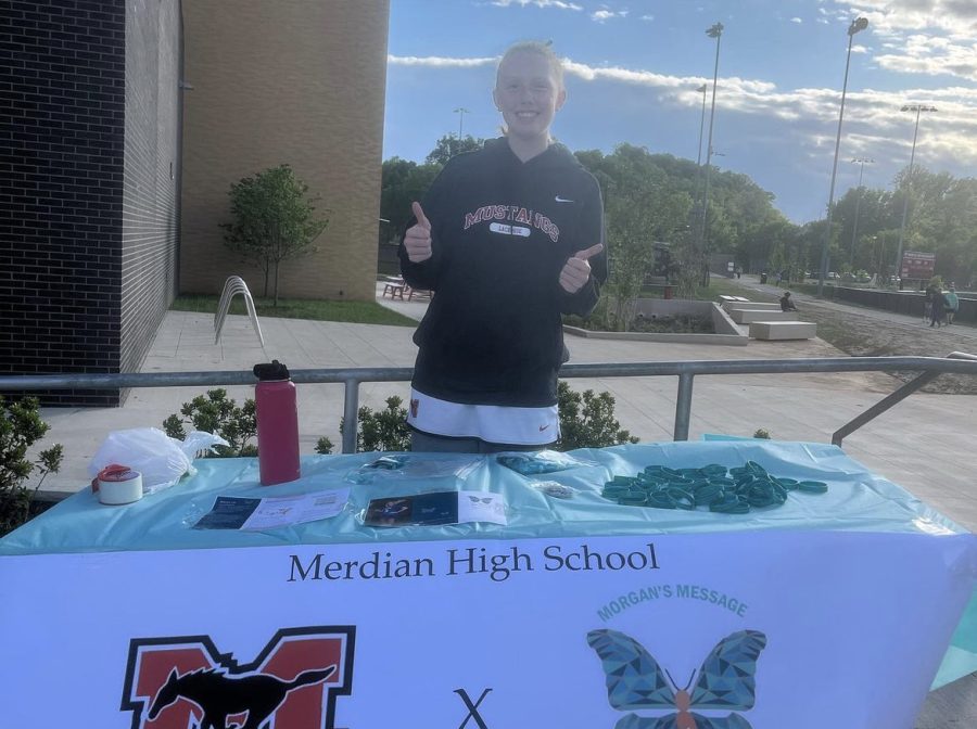 Sophomore Ally Campbell poses behind the Morgan’s Message information table at the clubs first dedication game. At dedication games, the club raises awareness for athlete mental health by educating spectators and involving the players in their pursuit. (Photo via @meridian_morgansmessage)