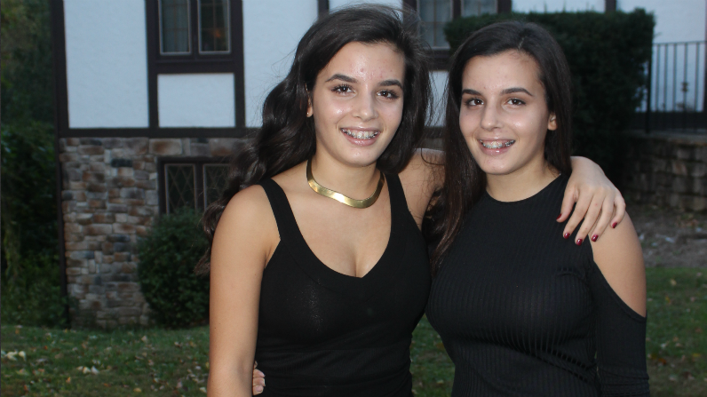 Identical twins in black dresses pose in front of a house.