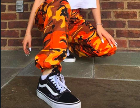A girl crouches with her feet towards the camera and her head not visible in front of a brick wall wearing a tee shirt and bright orange camouflage pants.