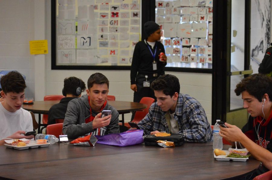 Four sophomore boys sit at a lunch table on their phones.