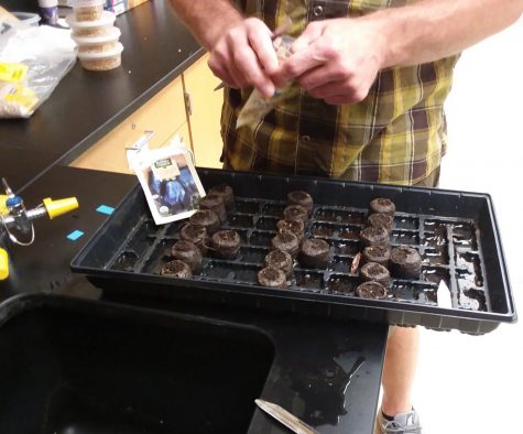 Mr. Perkins opens a bag of seeds. On the counter in front of him is a tray of small, environmentally-friendly soil pots. An opened seed bag rests inside the tray as well.
