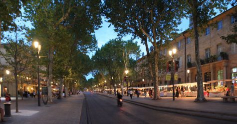 A street in Aix-en-Provence at early evening – it’s lined by shops and street lamps, and people walk about.