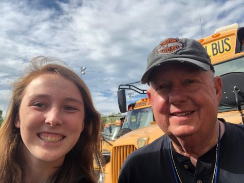 Two people smile for a selfie in front of a school bus. 