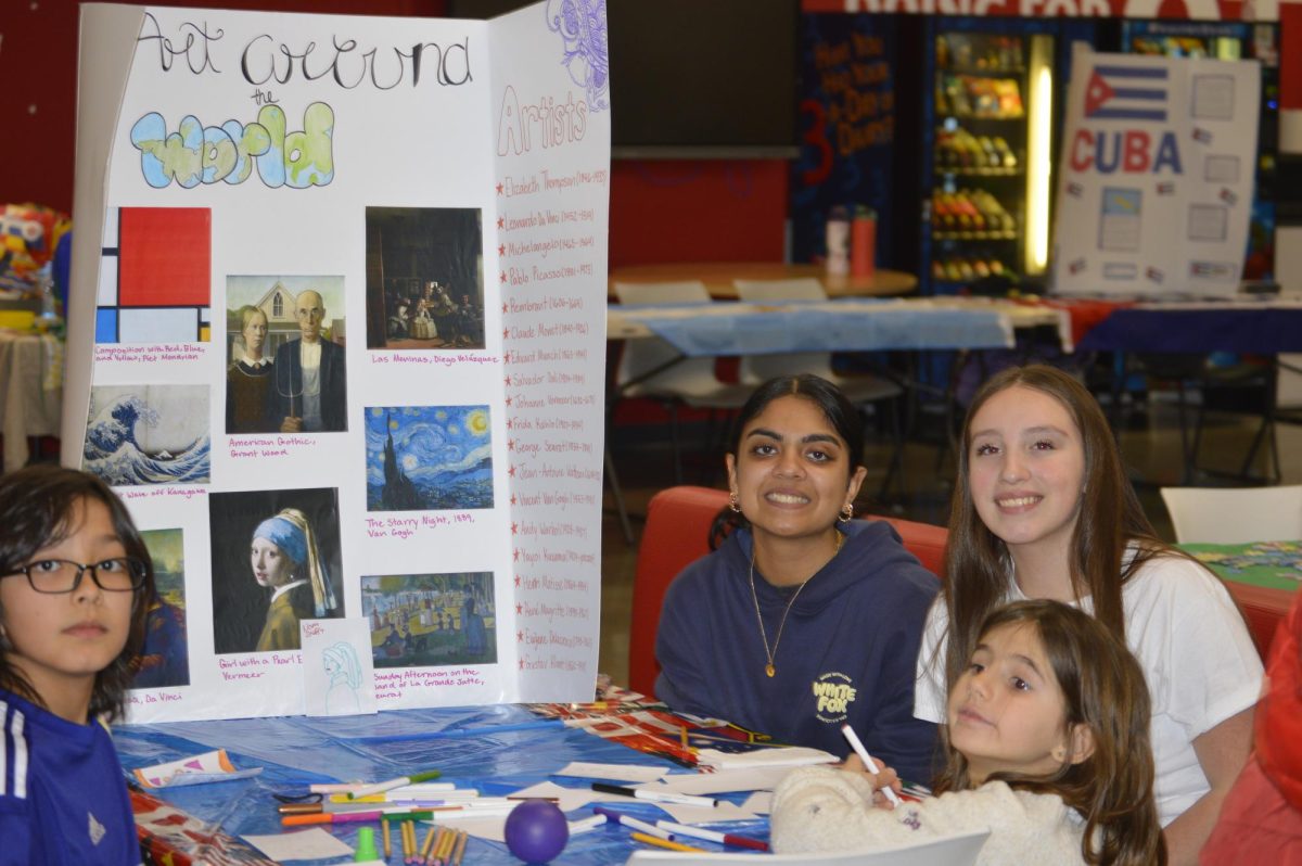 Freshmen Aditi Narain and Andrea Banuelos Valenzuela pose at their Art Around the World poster, as two kids color. (Photo by Layla Kozbelt)